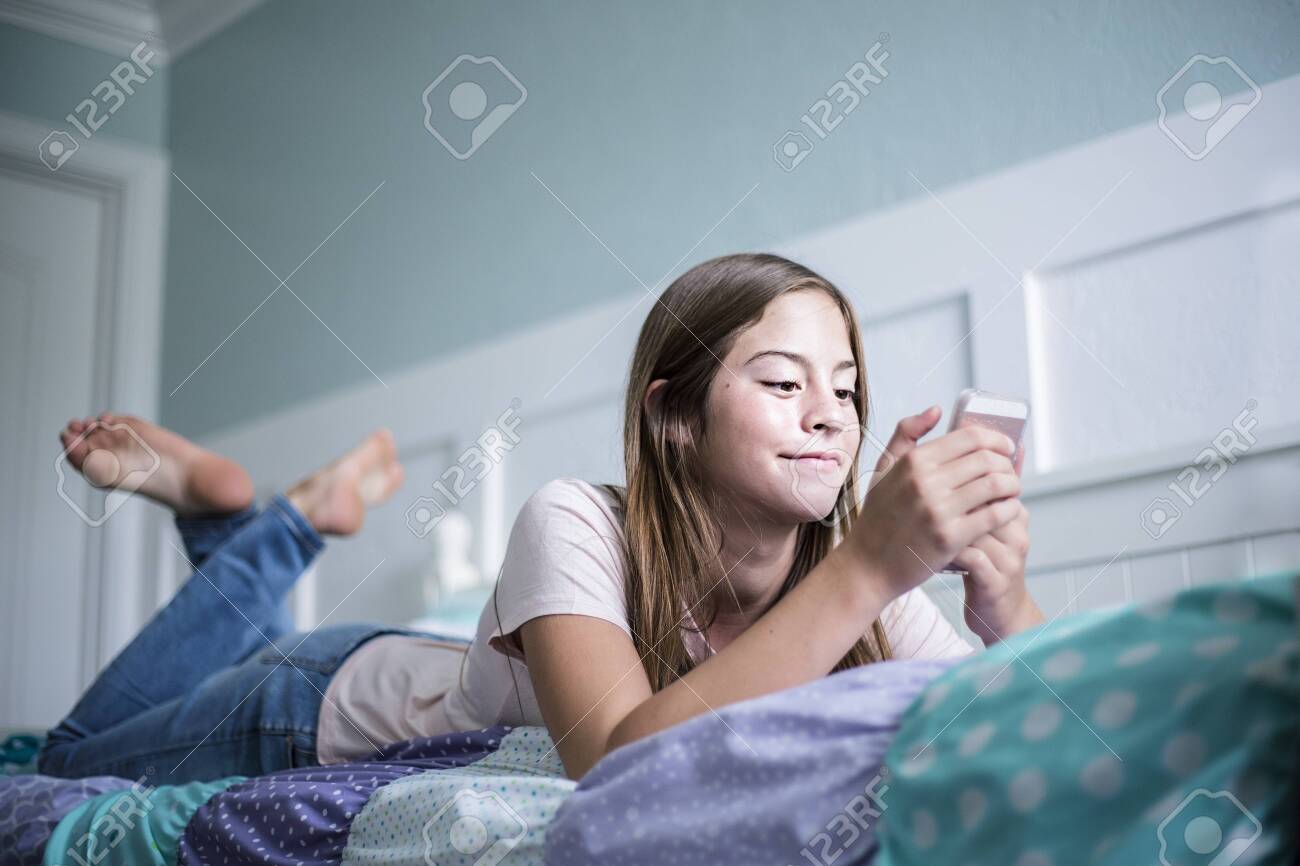 Pre-adolescent Teen Girl Texting On A Smartphone Lying In Bed At Home.  Candid Indoor Photo WithFocus On The Foreground And Copy Space Фотография,  картинки, изображения и сток-фотография без роялти. Image 142910709