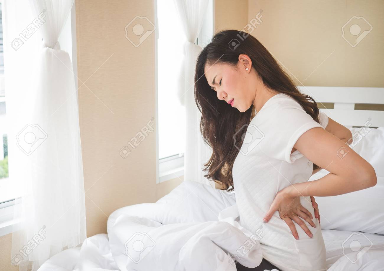 Young woman having back ache sitting on white bed. - 87676242 Young woman having back ache sitting on white bed. - 87676242