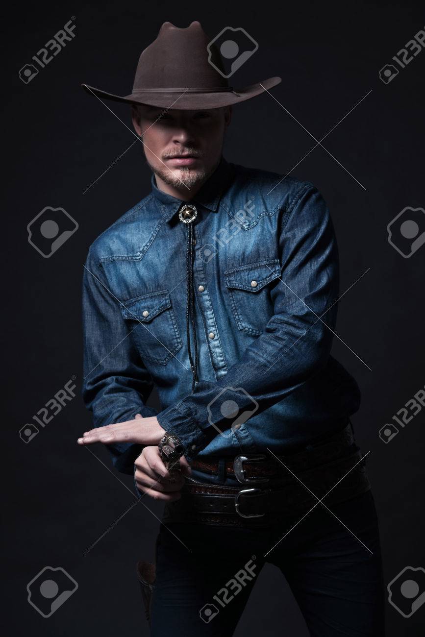 現代ファッション カウボーイ 茶色の帽子とシャツ ブルー ジーンズを着用します 彼の銃を引っ張る ブロンドの髪とひげ スタジオ ショット黒に対して の写真素材 画像素材 Image