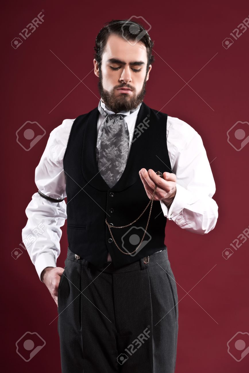 レトロな 1900年ビクトリア朝のファッション黒のジレとグレーのネクタイを身に着けてひげの男 懐中時計を保持しています 赤い壁を撮影スタジオ の写真素材 画像素材 Image