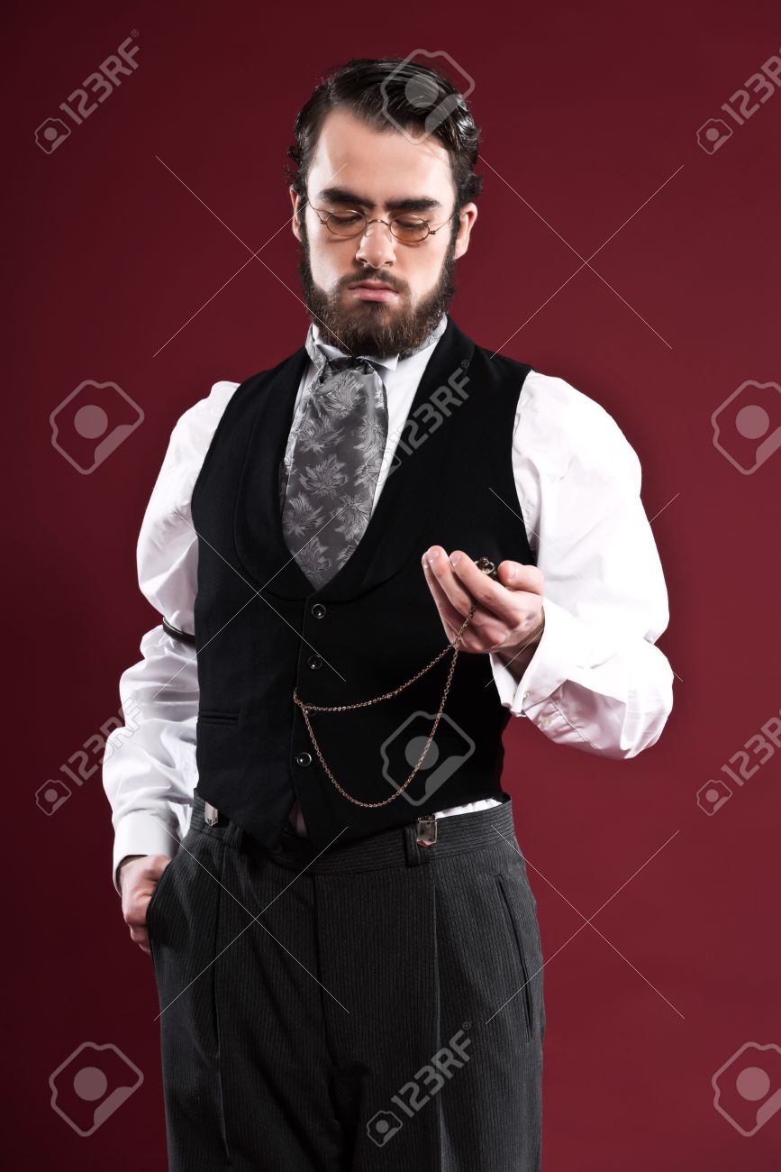 レトロな 1900年ビクトリア朝のファッション男ひげ黒ジレ グレー ネクタイとメガネを着用します 懐中時計を保持しています 赤い壁に撮影スタジオ の写真素材 画像素材 Image