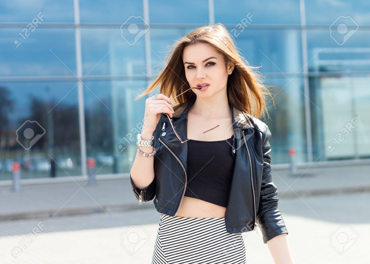 女の子モデル屋外のポートレート ガラス革のジャケットとスカートを保持しています 魅力のスタイリッシュな絶世の美人 若いファッション女性 の長い茶色の髪の摩耗のクールな服とサングラス ガラスの背景の建物します の写真素材 画像素材 Image