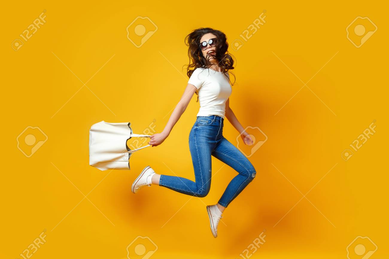 Beautiful young woman in sunglasses, white shirt, blue jeans posing, jumping with bag on the yellow background - 95851549 Beautiful young woman in sunglasses, white shirt, blue jeans posing, jumping with bag on the yellow background - 95851549