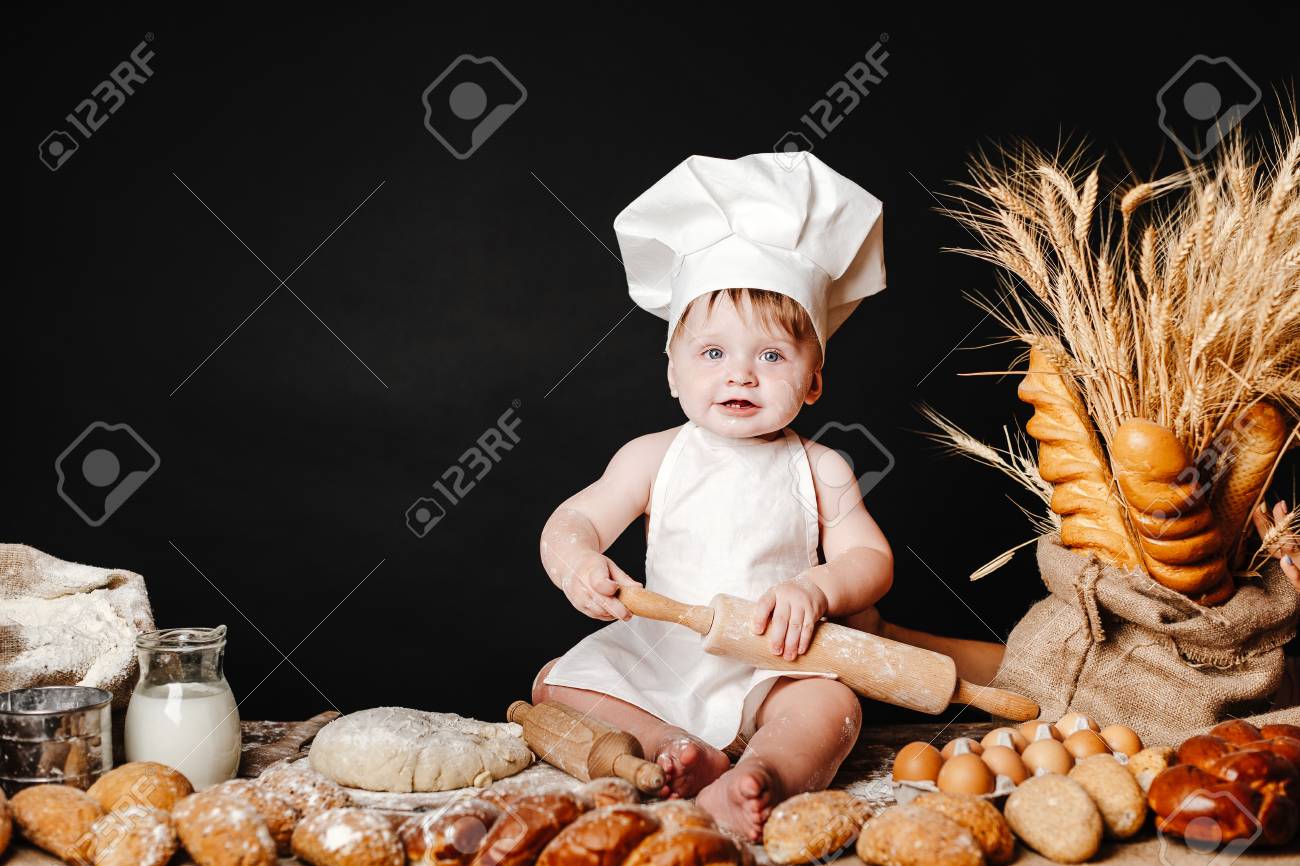 Charming toddler baby in hat of cook and apron sitting on table with bread loaves and cooking ingredients laughing happily - 106222740 Charming toddler baby in hat of cook and apron sitting on table with bread loaves and cooking ingredients laughing happily - 106222740