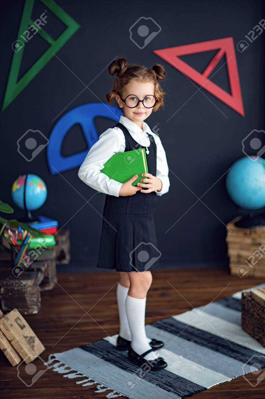 Clever little girl in school uniform and glasses smiling and looking at camera while holding green textbook before studies - 132323181 Clever little girl in school uniform and glasses smiling and looking at camera while holding green textbook before studies - 132323181
