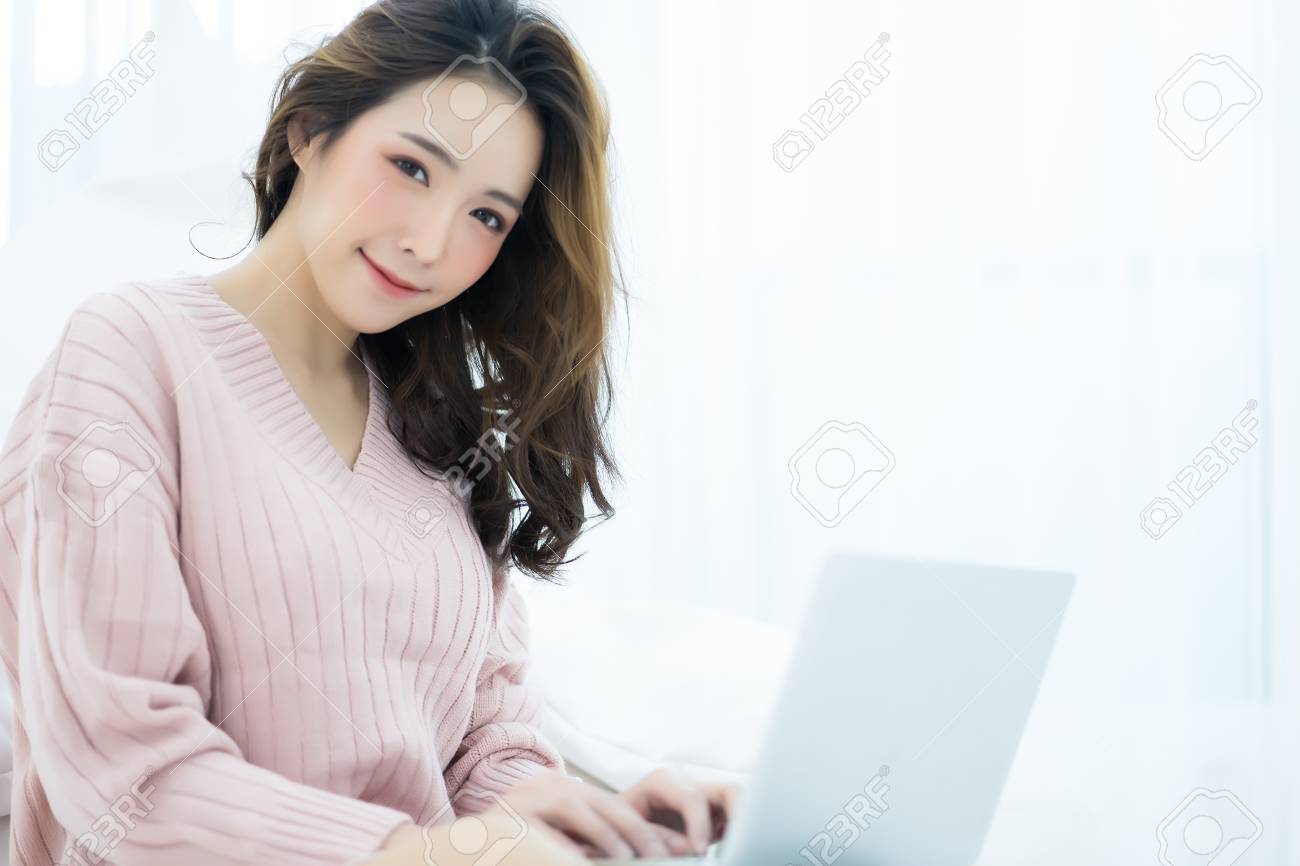 Beautiful young Asian woman working on a laptop on a laptop sitting on bed in the house in the winter looking at camera. Woman lifestyle Concept - 104673790 Beautiful young Asian woman working on a laptop on a laptop sitting on bed in the house in the winter looking at camera. Woman lifestyle Concept - 104673790