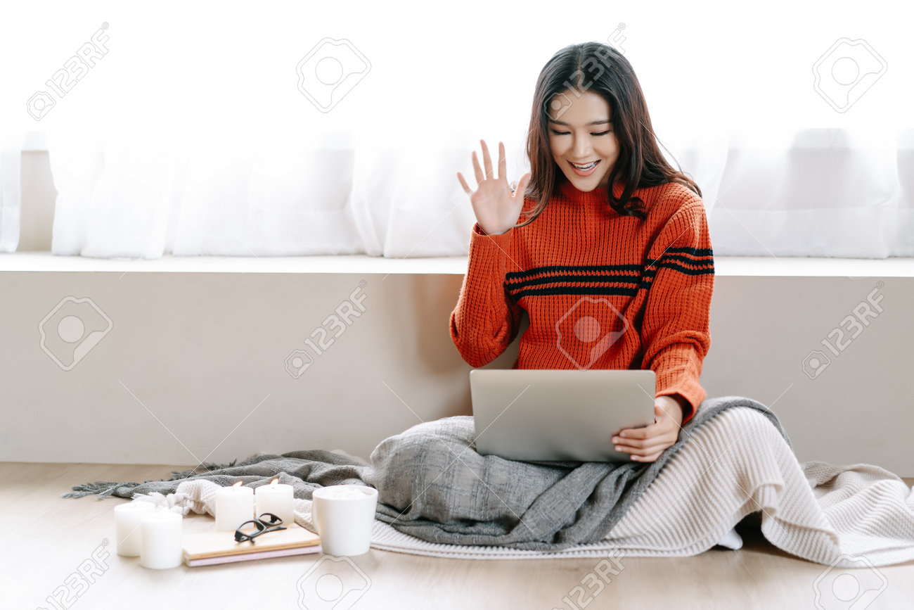 Portrait of young Asian woman smiling friendly using laptop computer have online video call broadcast meet bloggers say hi while sitting on floor at home in winter. Cozy winter decorated with candles. - 162360798 Portrait of young Asian woman smiling friendly using laptop computer have online video call broadcast meet bloggers say hi while sitting on floor at home in winter. Cozy winter decorated with candles. - 162360798