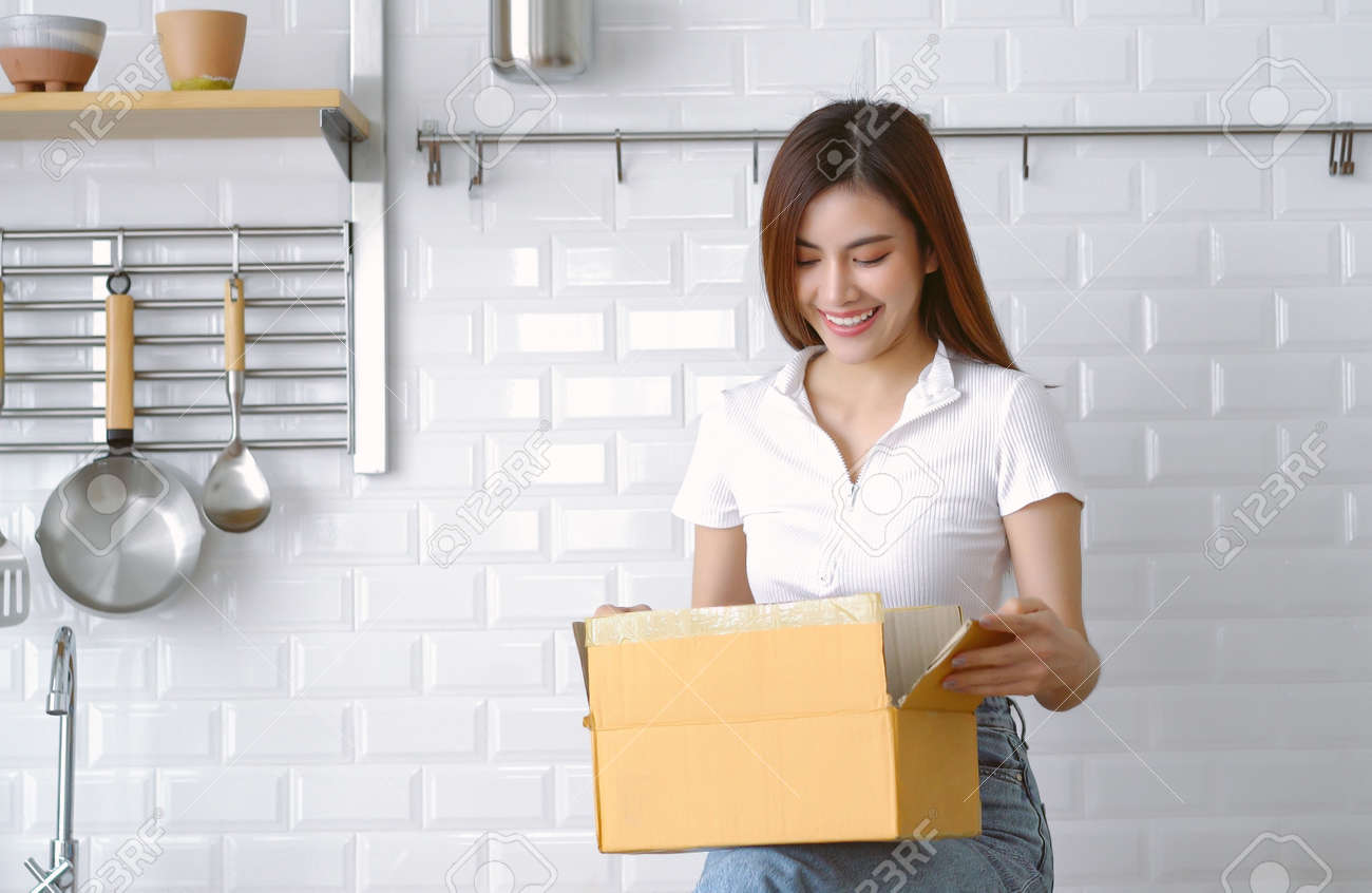 Una hermosa clienta asiática recibe un paquete de envío postal en casa. chica japonesa abriendo una caja de cartón mientras está sentada en la encimera de una cocina moderna blanca. orden de compra en línea, concepto de entrega - 189296196 Una hermosa clienta asiática recibe un paquete de envío postal en casa. chica japonesa abriendo una caja de cartón mientras está sentada en la encimera de una cocina moderna blanca. orden de compra en línea, concepto de entrega - 189296196