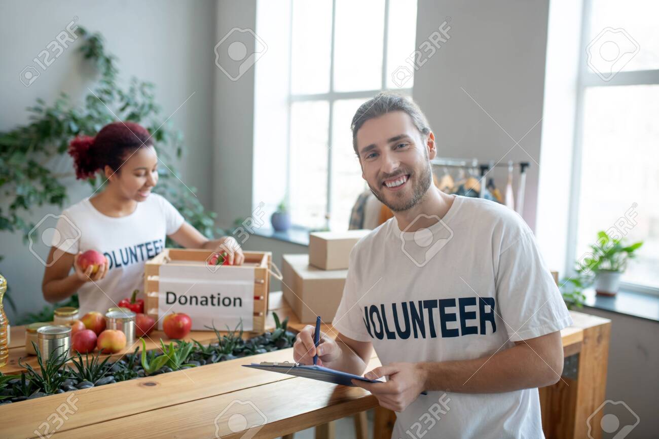 Food aid. Young man in front of the table writing notes, the girl at the table putting apples in a charity box, both of them in a good mood. - 140078164 Food aid. Young man in front of the table writing notes, the girl at the table putting apples in a charity box, both of them in a good mood. - 140078164