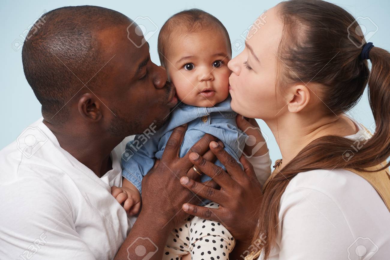 Portrait Of Happy Multiethnic Family White Mother Black Father And Their Adorable Little Baby Daughter Against Pale Blue Background Both Parents Kissing Their Daughter One Cheek For Each Close Up Stock Photo