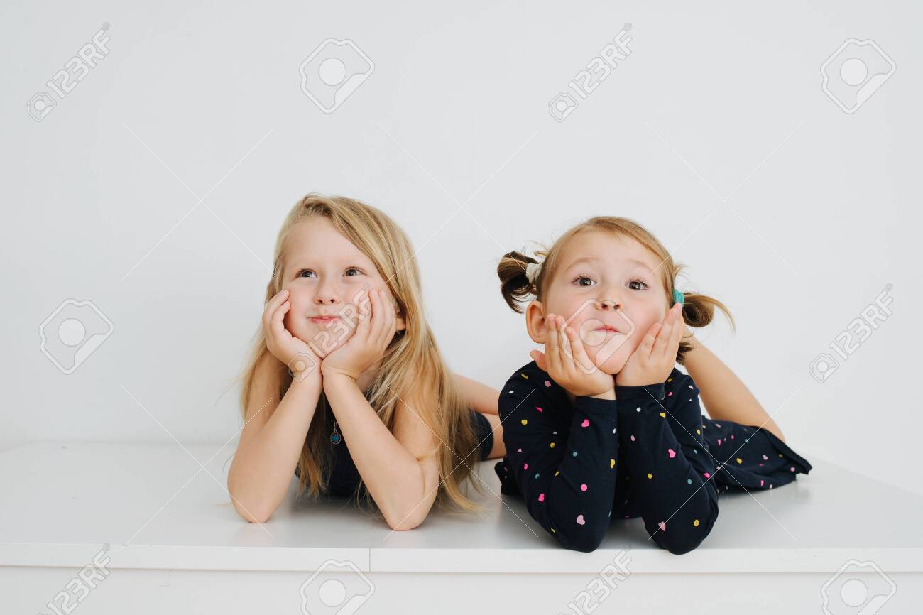 Adorable little barefoot siblings lying on a table with their feet up. Over white wall. Inflating their cheeks, leaning on hands, making cute faces for the camera. Both in nightware. - 143650456 Adorable little barefoot siblings lying on a table with their feet up. Over white wall. Inflating their cheeks, leaning on hands, making cute faces for the camera. Both in nightware. - 143650456